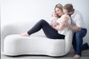 A candid studio photo portrait of a husband and wife embracing their newborn baby together on a modern white couch. By Mike Streeter Photography, in Milton, Ontario, Canada.