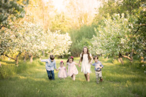 A candid photo portrait of 5 young children all holding hands while running towards the camera through an apple orchard.By Mike Streeter Photography in Milton, Ontario, Canada.